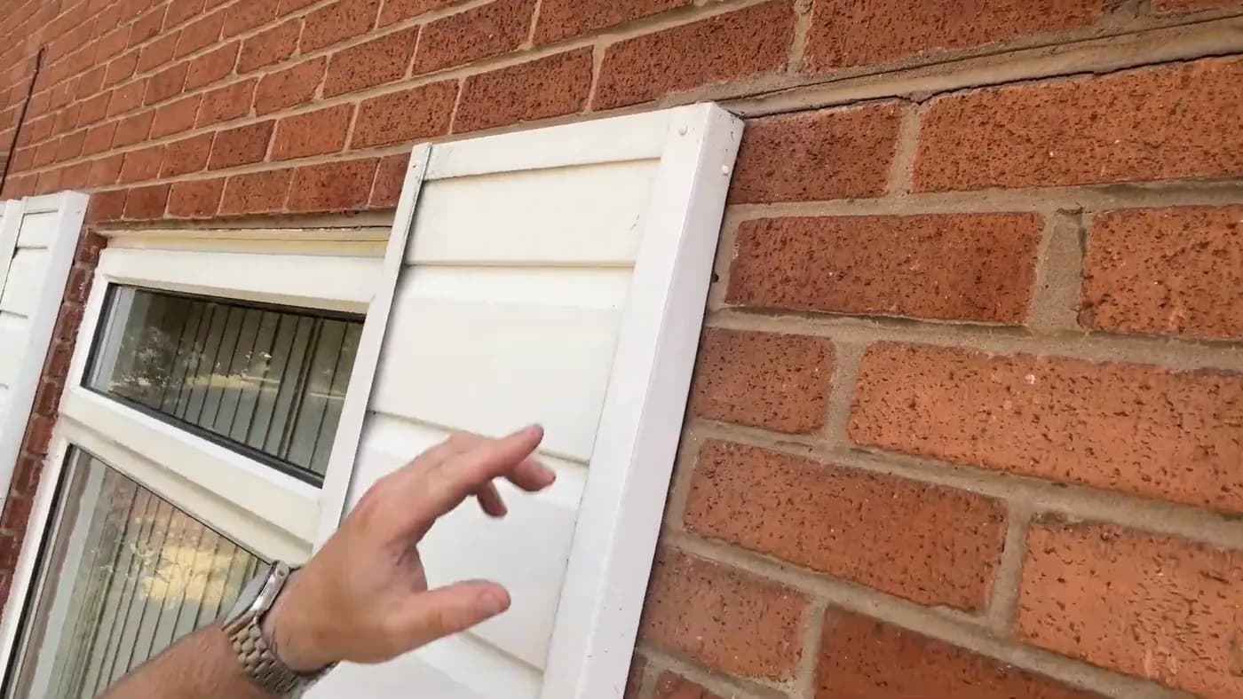 A hand pointing at the junction between a window frame and the surrounding brickwork.