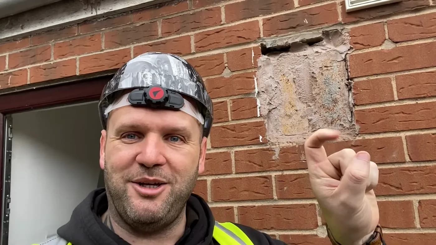 John pointing at a crude mortar patch spread across brickwork beside a doorway.