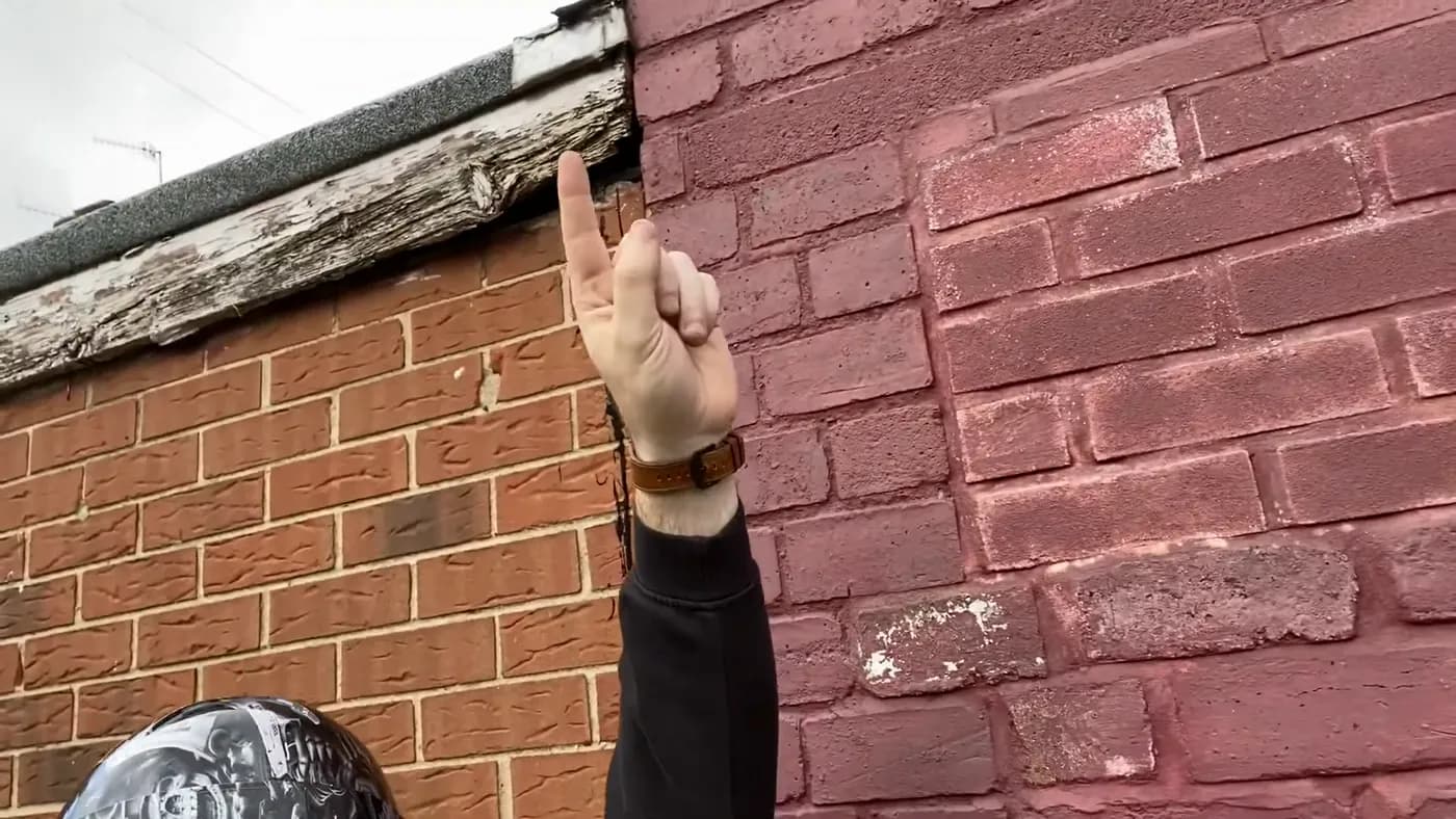 John pointing up at a rotten timber fascia board above painted brickwork.