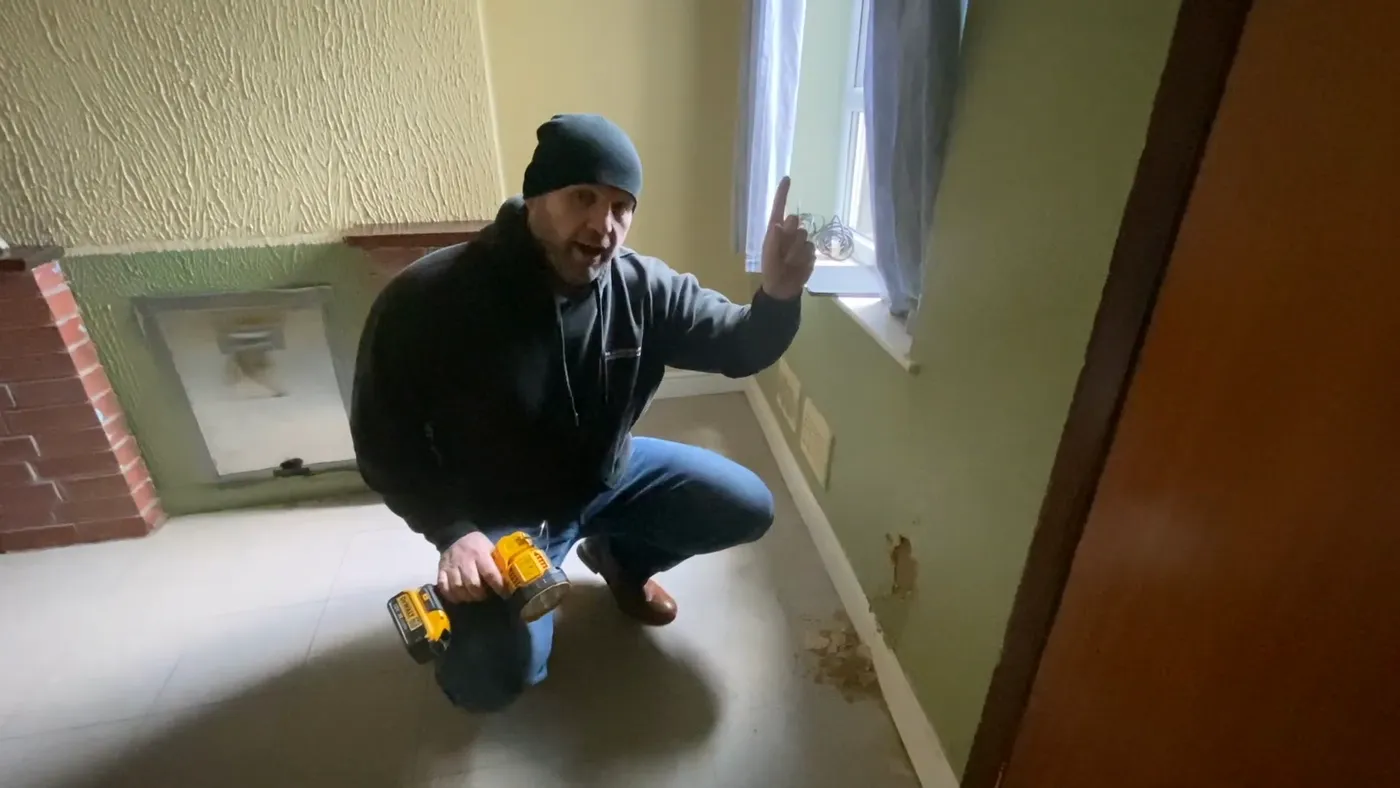 John kneeling at the base of a wall taking a reading with a hand-held damp meter.