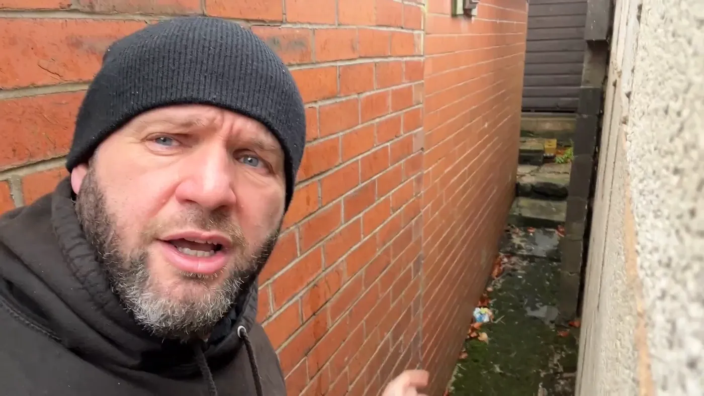 John pointing along an external brick wall at ground level where surface water is running against the house with no drainage.