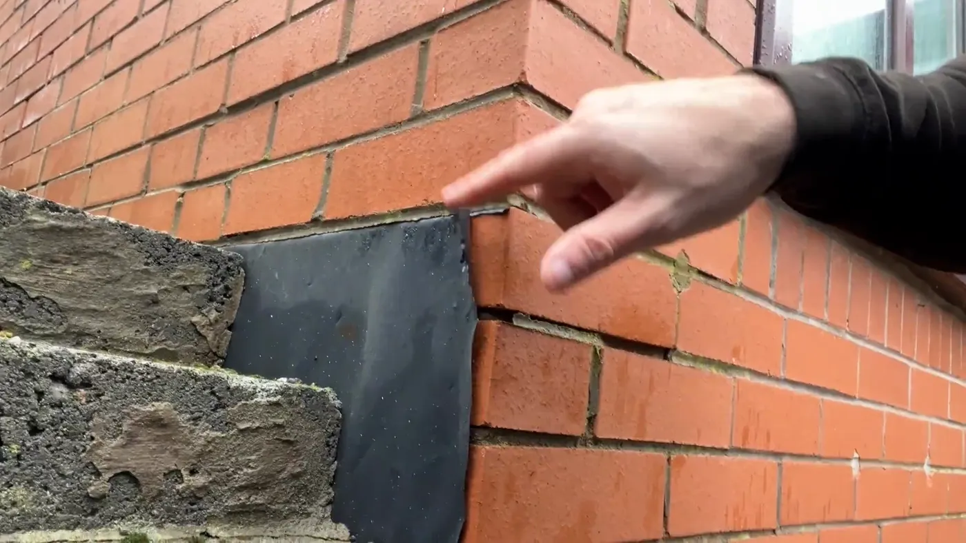 A hand pointing at a thin sheet of black DPM squeezed between external brickwork and steps built up against the house.